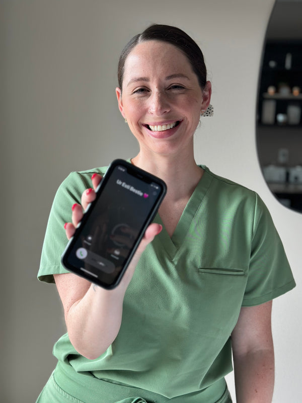 Katie A., Licensed Esthetician at Young & Bloom Skin in Saratoga Springs, playfully holding up a phone to the camera inviting clients to book an appointment.