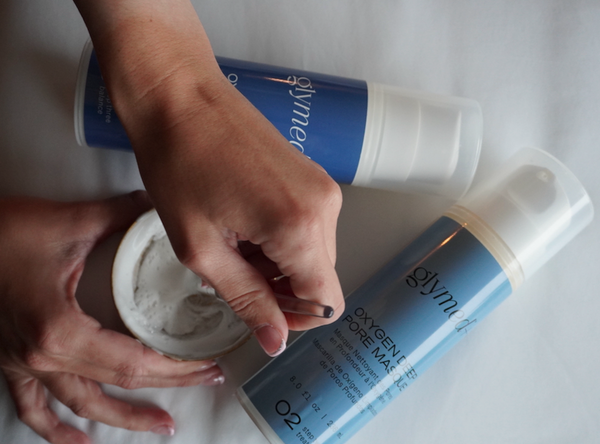A close-up of hands applying or mixing a white cream from a small white jar into a tube of blue Glymulin Oxygen-Pure Pore-Makeup on a white fabric background.